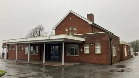 Thorncliffe Crematorium, which is a brick building with a white flat roof above a covered walkway entrance. The ground is wet from rain.