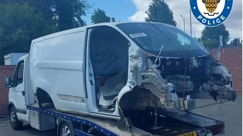 West Midlands Police A white van is loaded on the back of a white truck. There is a brick wall in front and to the side of the vehicles, which appear to be in a car park of compound.