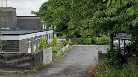 A car park and some grey brick buildings surrounded by trees. On the right a white sign reads: Knowle Manor