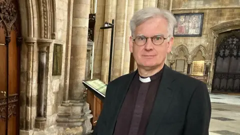 A man with light grey hair stands inside a medieval church. He is wearing round glasses, a dark blazer, dark-purple shirt and white dog collar.