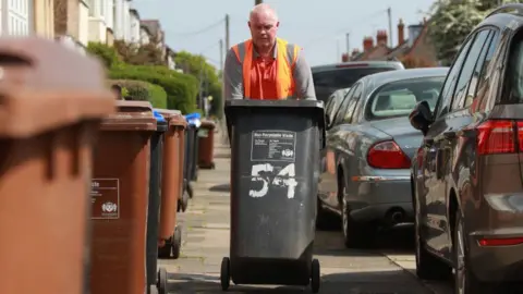 Getty Images A man wearing a high vis orange shirt is pushing a black wheelie bin towards the camera. He is on a pavement, lined on one side by more wheelie bins and the other by cars.