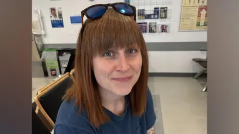 Sam Naylor smiling up at the camera. She has sunglasses on her forehead and red hair with a fringe.