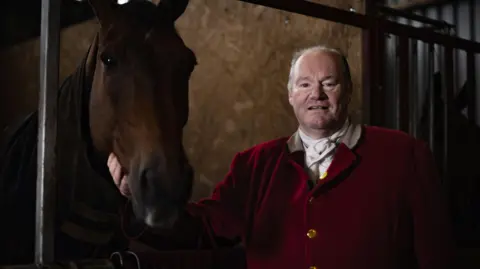 BBC A huntsman successful  reddish  overgarment   looks astatine  the camera with his horse