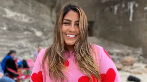Viviana is standing on a sandy beach in front of a concrete wall, wearing a pink sweater with red heart patterns. Several people sit on folding chairs and blankets in the blurred background.