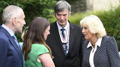 PA Media A man in a navy blue suit and colourful tie and a woman with long brown hair and a green dress greet writer Sir Ian Rankin, who wears a black suit with a navy blue tie and official medal, and Queen Camilla, wearing a black and white polka-dot dress with a white collar.