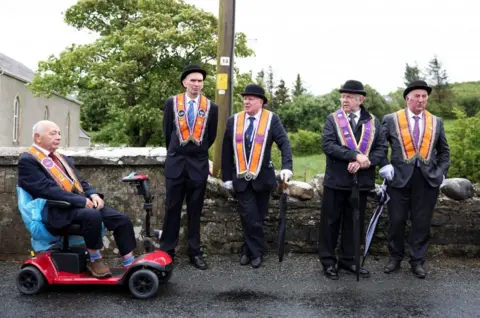 Five men are side by side in front of a stone wall. Each of them are wearing orange and purple sashes around their necks, dark blazers and bowler hats. 
The man on the far left is sitting in a red mobility scooter