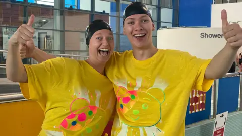 BBC Gemma and Luca swim with their mouths open looking at the camera. They're by a pool with the showers in the background. They've both got their Children in Need swim caps and bright yellow and baggy tops with Pudsey's face on them.