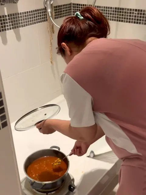 A woman in a pink and white outfit stands in a bathroom with white tiles and black mosaic accents, holding a pot lid and stirring a pot of soup placed on the floor. Their hair is tied back with a green hair tie.