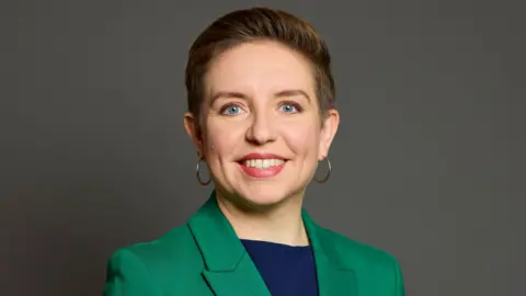 UK Parliament Green Party leader Carla Denyer faces the camera in an official Parliament photograph. She is wearing a dark blue top and green jacket and has short hair