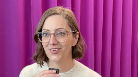 A woman, Jackie Jantos, with glasses and a white top, in front of a purple curtain.
