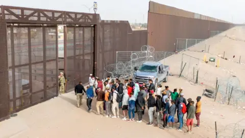 An aerial view of a group of people waiting to be processed at the US Mexico border. At the border is a high metal fence stretching into the distance.