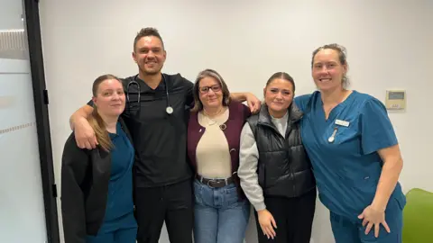 John Devine/BBC Five people are standing inside an office smiling at the camera. Three are wearing medical overalls.