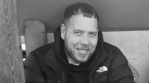A black and white picture of Michael Knowlson and his son Mason sitting beside each other in a wooden structure at a playpark, smiling at the camera.