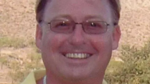 Cheshire Police A man smiling, wearing glasses and a yellow shirt with a silver necklace. 