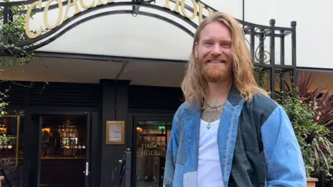 Sam is stood outside a pub, smiling down towards the camera, which is angled slightly lower than him. He has shoulder-length, strawberry blonde hair and beard. He is wearing a denim jacket with a white top underneath and two short necklaces. 