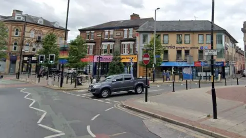 Google A view of the centre of Withington by the suburb's public library, with a car leaving one road to join another by a set of traffic lights with shops seen in the background. 