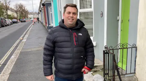 Roger Smith wearing a black North Face coat stands on a street outside a row of terrace houses. The door next to him is lime green.