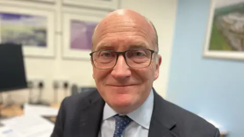 Nick Hulme smiles directly at the camera. He is wearing dark-framed glasses, a grey suit, white shirt and blue and white polka dot tie
