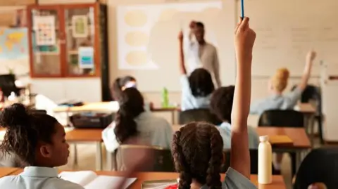 A female pupil raises a hand in a classroom full of children and a teacher.