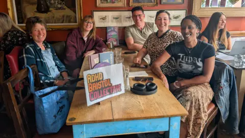 Nathalie Giaquinto Five adults are sat round a wooden table at a restaurant. On the table is a sign which reads 'ADHD Bristol Collective'