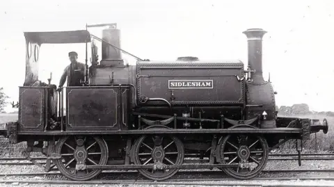 Manhood Wildlife & Heritage Group A historic photograph showing the steam locomotive Sidlesham standing on railway tracks. A railway worker stands on the locomotive, which has the name "SIDLESHAM" displayed on its side.