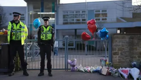 PA Media Two police officers wearing their uniform stood outside the metal school gates, with the school building behind them. An array of flowers and balloons are placed in a row by the gates.