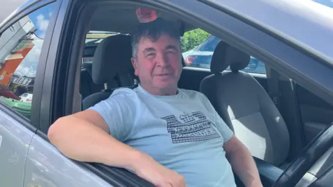 John Moreton is sitting inside his car. The window is down and he is looking at the camera, wearing a blue t-shirt. 