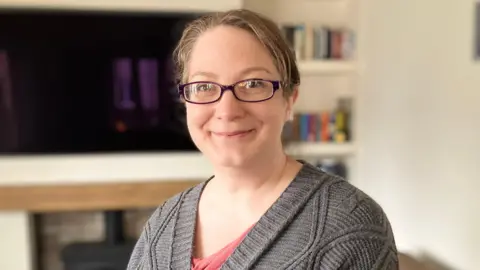 Hannah has light brown hair tied back and is wearing a pink top and grey cardigan. She wears black rectangular glasses