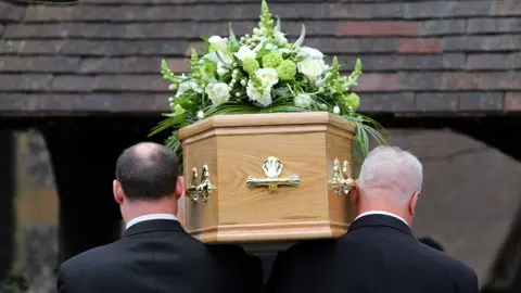 A view from behind of a coffin being carried into a building on the shoulders of pallbearers dressed in black suits.