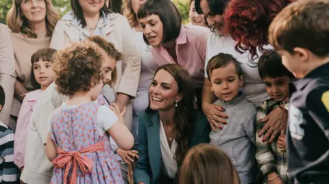 Royal Foundation Kate, smiling, wears and blue blazer. She crouches, surrounded by smiling women and children