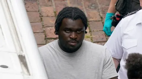 Anthony Esan is wearing a grey T-shirt and looking straightforward as he stands next a police officer.