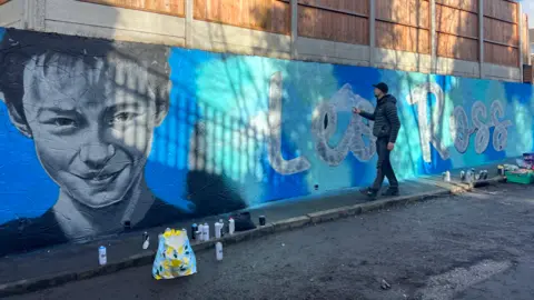 A man touches a wall on which a gigantic blue mural has been painted. On the left is Leo Ross's face, neck, and shoulders. On the right is his name, painted on the wall in white writing.
 