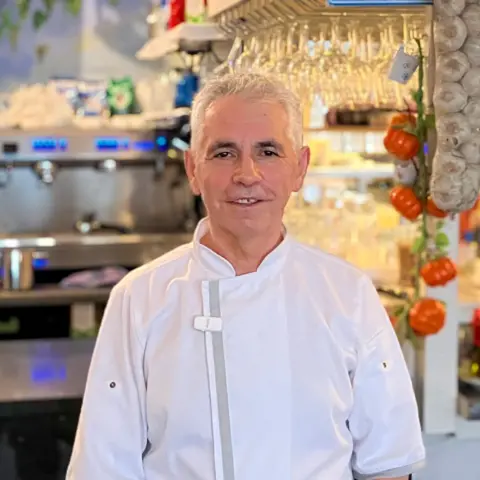 Petrit wearing a white chef’s jacket standing inside his restaurant, just in front of the kitchen and bar. Shelves behind him hold hanging utensils, spices, and kitchen equipment. Bottles of spirits and Greek decorations are arranged along the back wall. There is also an espresso machine visible in the background.
