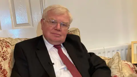 Dr Tim Brain, wearing a dark suit and red tie, sitting at home on a sofa.