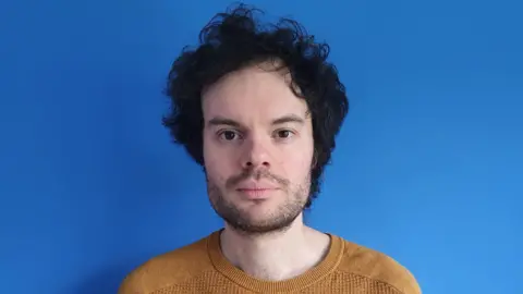 A man in a mustard yellow jumper with black stubble and thick black hair stands in front of a blue wall