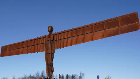 PA Media Antony Gormley's Angel of the North sculpture in Gateshead. The tall brown sculpture, which resembles a man with rectangular wings, stands tall over the wiry trees below.