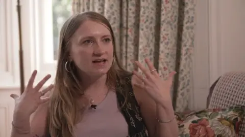 Marianne gesturing with her hands as she speaks. She has long blonde hair and has a nose piercing, and is wearing a pink short with a brown waistcoat.