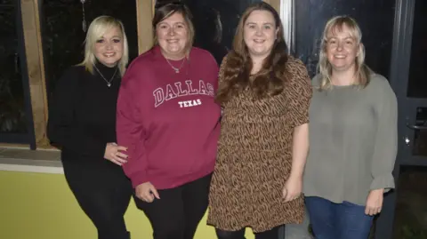 Frome Times Four women standing in a line and smiling to camera. From left, one has blond hair and black clothing, the second has a purple sweatshirt saying 'Dallas Texas', the third one is wearing a leopard-print long top and the last woman is in a grey top and jeans. They are in a club foyer-type space