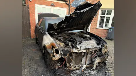Salma Yaqoob A photo of a burnt-out car with the bonnet up. It is parked on the drive outside a house.