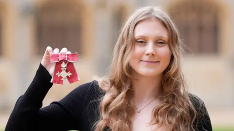 ParalympicsGB's Poppy Maskill with long blond hair wearing a black dress smiles as she holds up her Member of the Order of the British Empire medal