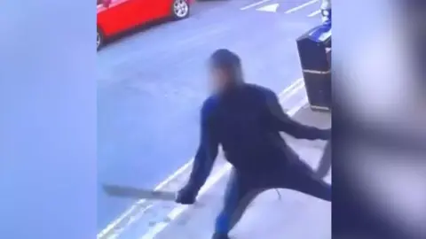 Anonymous A CCTV image of a man with a blurred face, wearing dark clothing, wielding a machete in his right hand