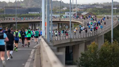 Great Scottish prowadził dużą grupę biegaczy biorących udział w wyścigu na podwyższonej jezdni, z jasnym sprzętem sportowym i scenerią miasta w tle.