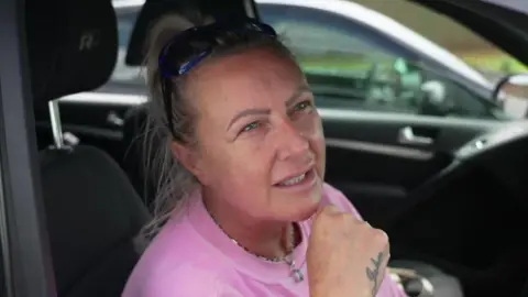 Julie Pemberton, who has blonde hair tied back into a bun and wears a pink t-shirt, speaks to the camera out from the driver's  side window of her car