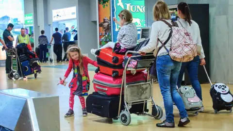 A family with two small children push a luggage trolley pilled with red and pink suitcases through an airport . One of the chidlren is sitting on top of  a suitcase and being pushed  through the airport by a parent