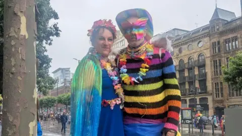 Kit Taylor/BBC Two people wearing very bright colours and headwear stand side by side against the backdrop of a wet city centre street. The person on the left is wearing a rainbow-coloured cape and a blue dress, while the figure on the right is wearing a rainbow-themed hat, butterfly mask and jumper.