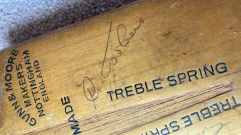 Richard Bond A close-up image of a section of an old cricket bat, a light brown in colour with black text printed on it. To the left of frame, some text sideways-up as seen in the image reads "Gunn & Moore, Makers, Nottingham, England". The word "Made" is also visible below this, and to the right of frame "Treble Spring" written lengthways. In the centre of the frame is a written signature.