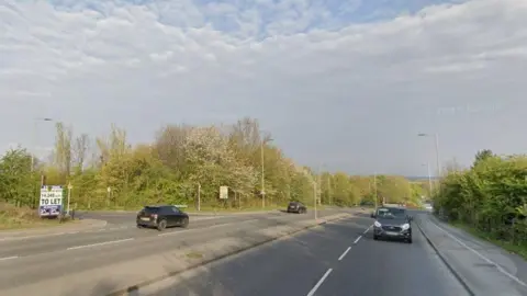 A road with cars driving down both sides. There are several street lights and greenery on the roadside. 