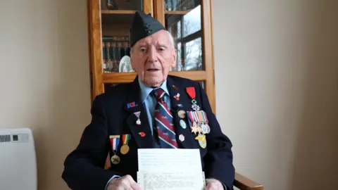 BBC An elderly man in a blue suit with lots of military medals pinned to his jacket. He  wears a blue military cap and holds a telegram in his hands. He is sitting in a chair in the room of a house, with a cabinet behind him and a radiator to the left.