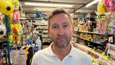 Sam Read/BBC A picture of a man in front of the market stall he owns. It sells a wide variety of goods, and he is wearing a white polo shirt. He has short hair and a goattee beard.