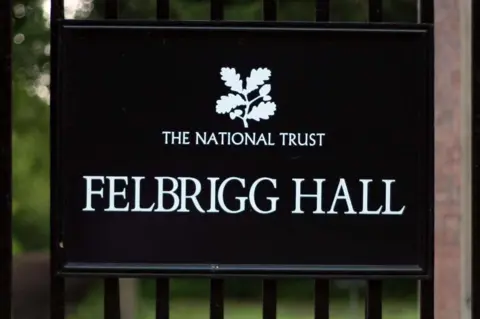 PA Media A black rectangular sign mounted on metal gate railings features a white oak leaf logo and "THE NATIONAL TRUST" at the top, with the words "FELBRIGG HALL" written below it in a large, clean, white serif font.